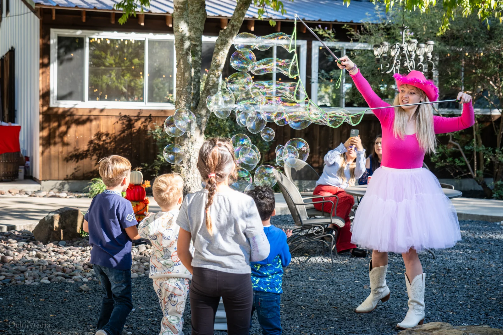 Children chasing bubbles around a performer in a pink tutu at a backyard party