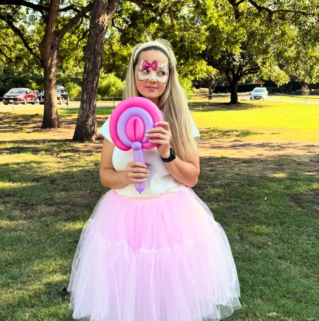 A performer in a pink tutu holding a twisted balloon lollipop in a park