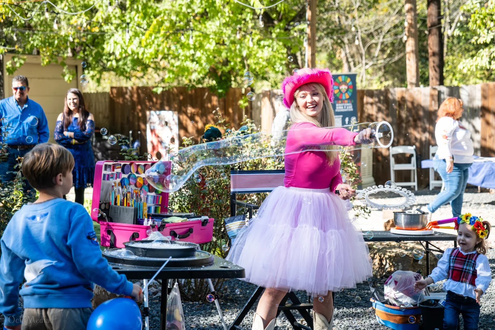 A performer making giant bubbles beside a face painting station as children watch